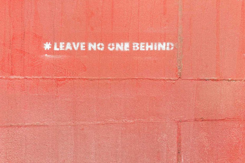 Foto van een rode muur met wit stencil opschrift: leave no one behind